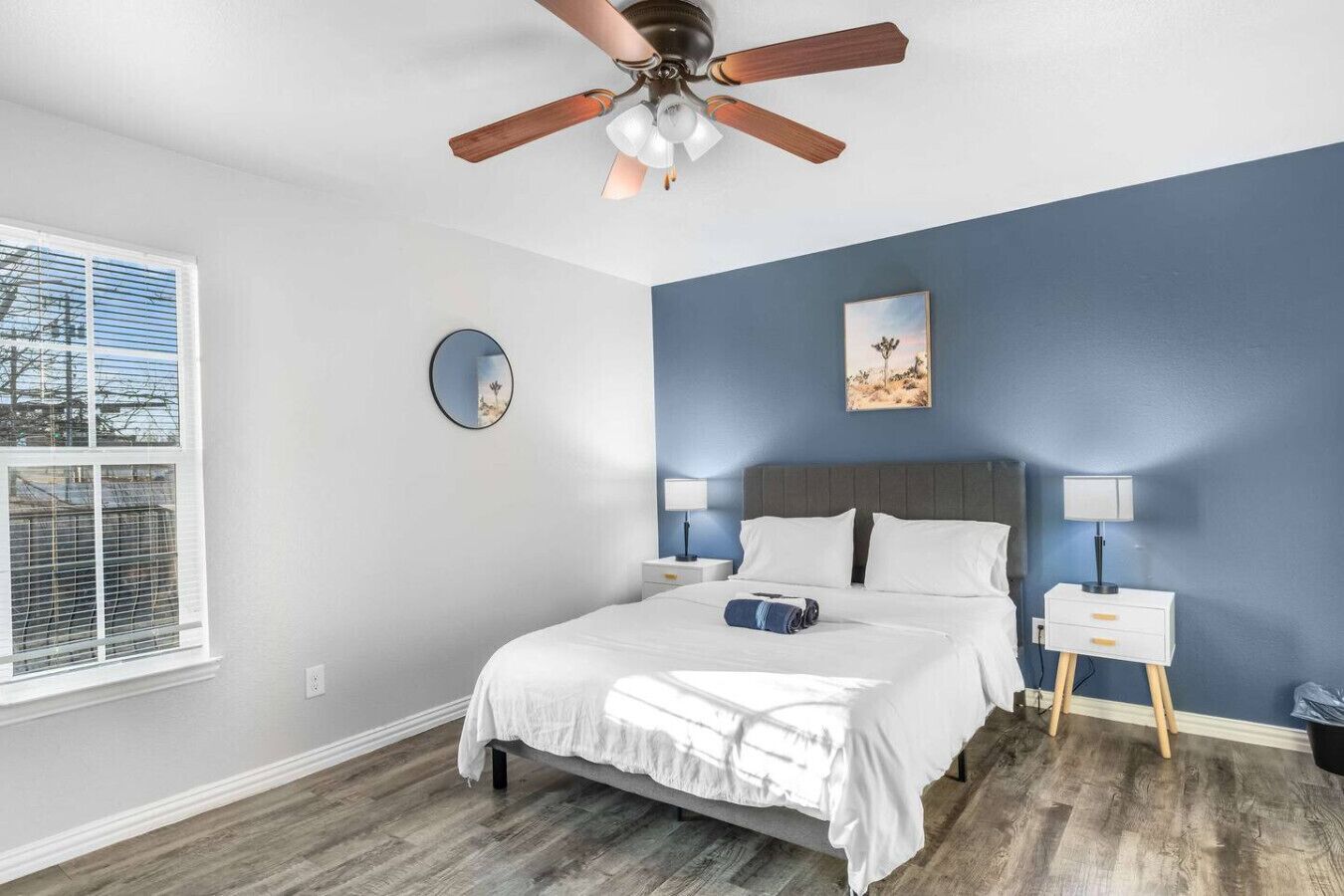 A bedroom with a bed, a ceiling fan, a window with blinds, a bedside table with a lamp, and a wall with a painting.