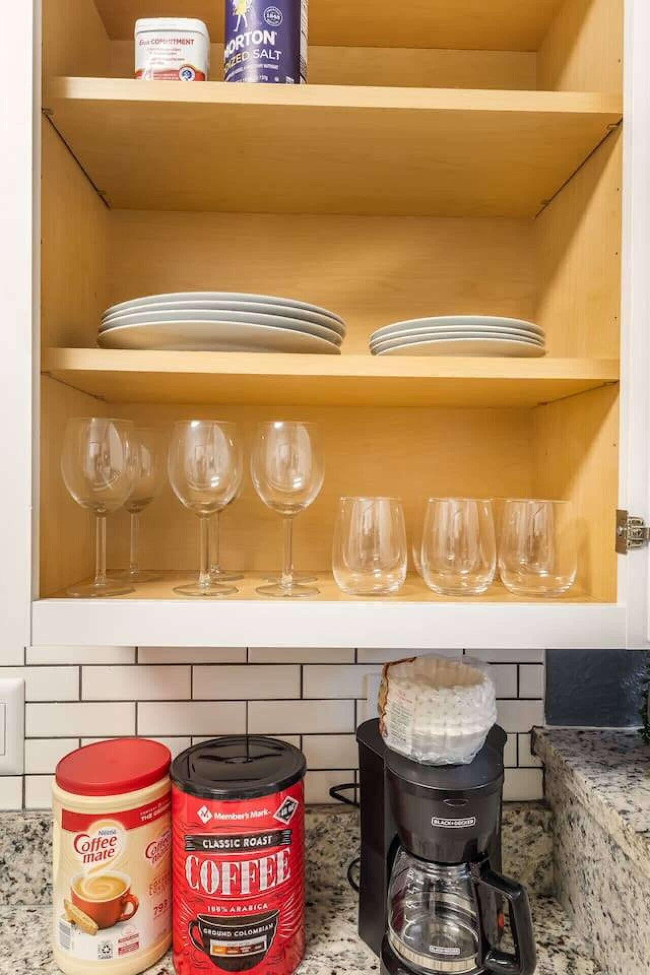 A kitchen with a wooden cabinet shelf holding plates and glasses, a coffee maker, and two coffee containers.