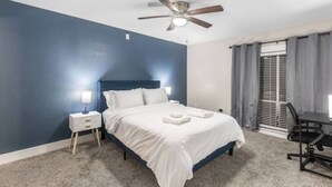 A bedroom with a bed, a desk, a chair, a ceiling fan, and a window with curtains.
