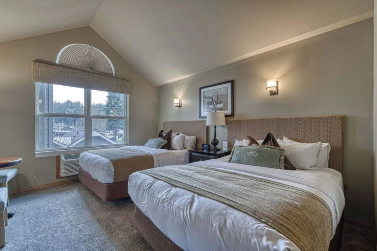 A hotel room with two beds, a desk, a chair, a window with a view, and a framed picture on the wall.
