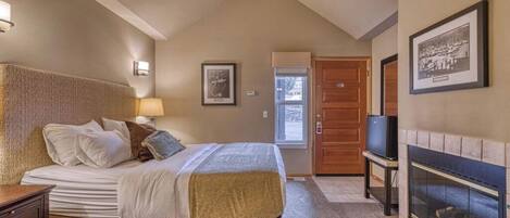 A hotel room with a large bed, a television, a fireplace, and a door leading to another room.