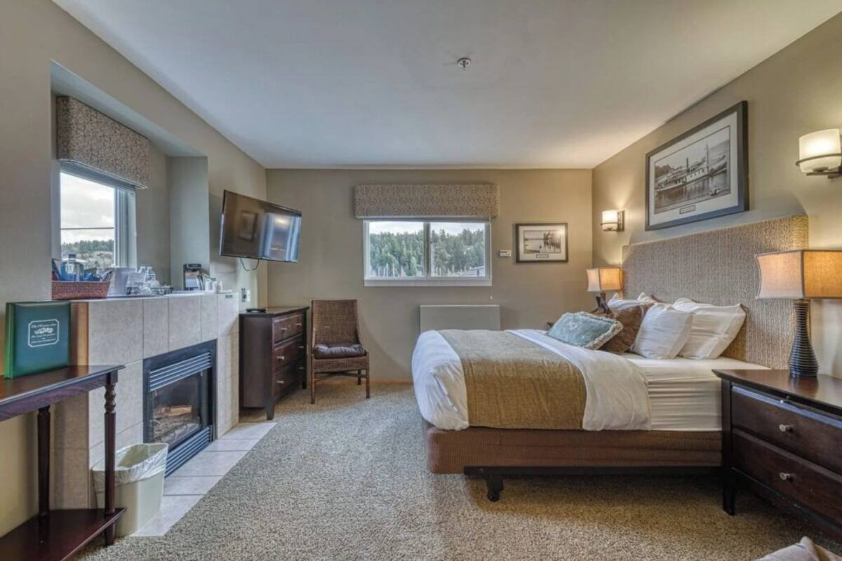 A bedroom with a bed, bedside tables, a TV, a fireplace, and a window with a view of trees.