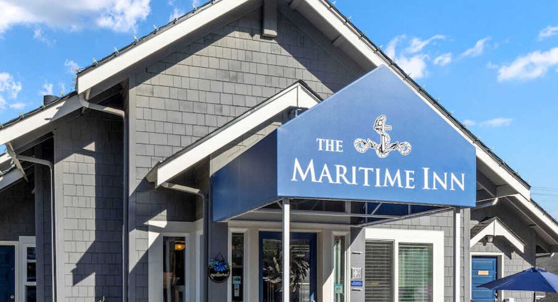 The exterior of The Maritime Inn, featuring a prominent sign with nautical-themed logo, a modern architectural design with grey brickwork, and a clear blue sky in the background.