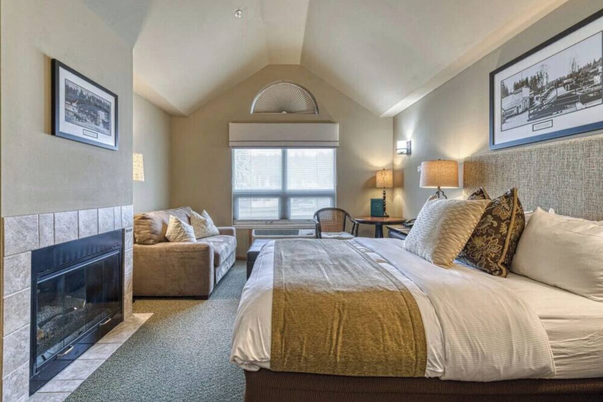 A hotel room with a fireplace, a bed with multiple pillows, a sofa, a desk with a lamp, and framed pictures on the walls.