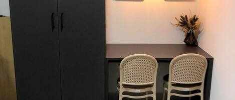 Una habitación con un armario oscuro, un escritorio con dos sillas de mimbre, una lámpara de pared y un jarrón pequeño con plantas secas.