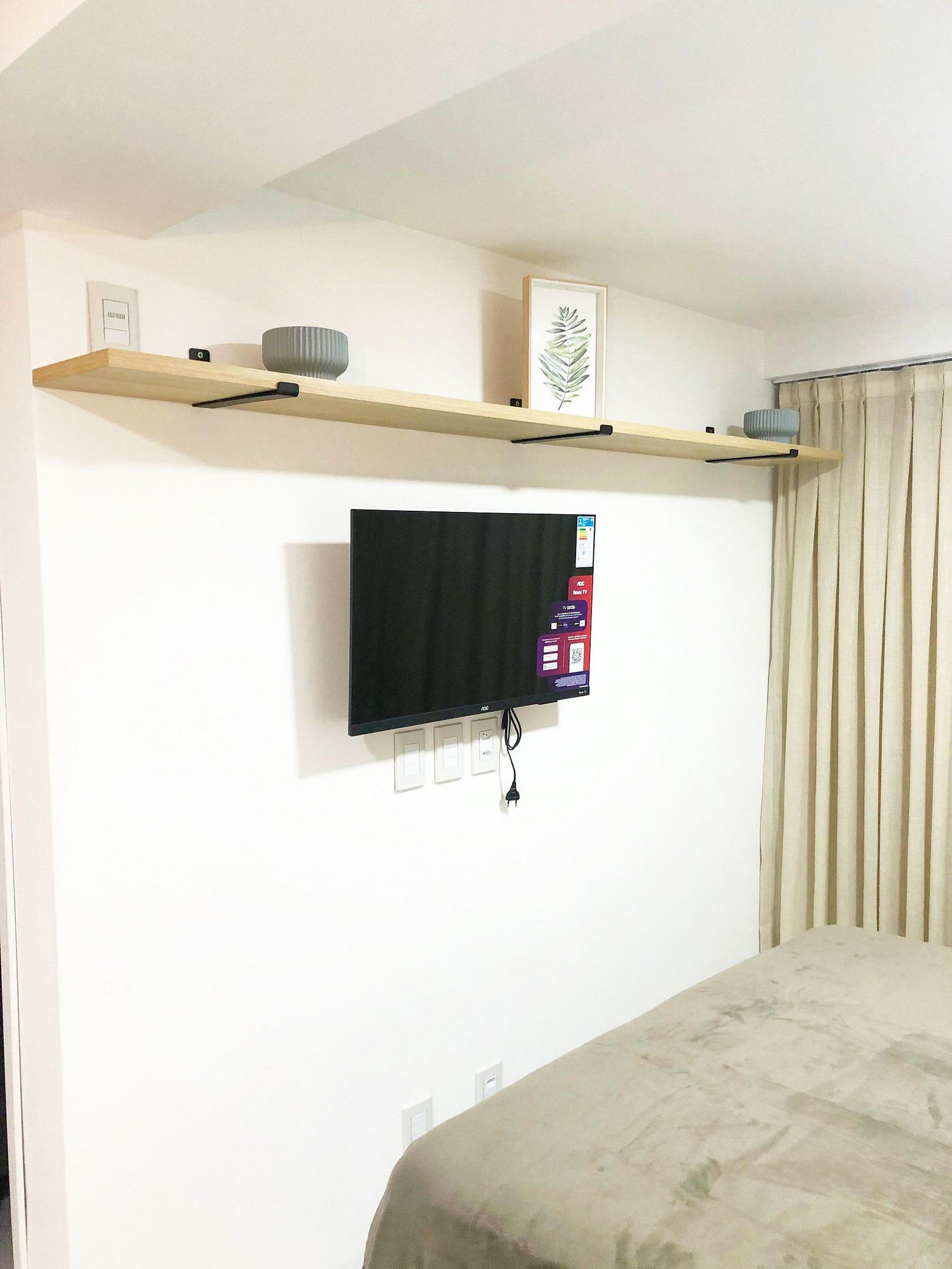 Una habitación con televisor de pared, estante de madera con objetos decorativos y cama con cubierta color beige.