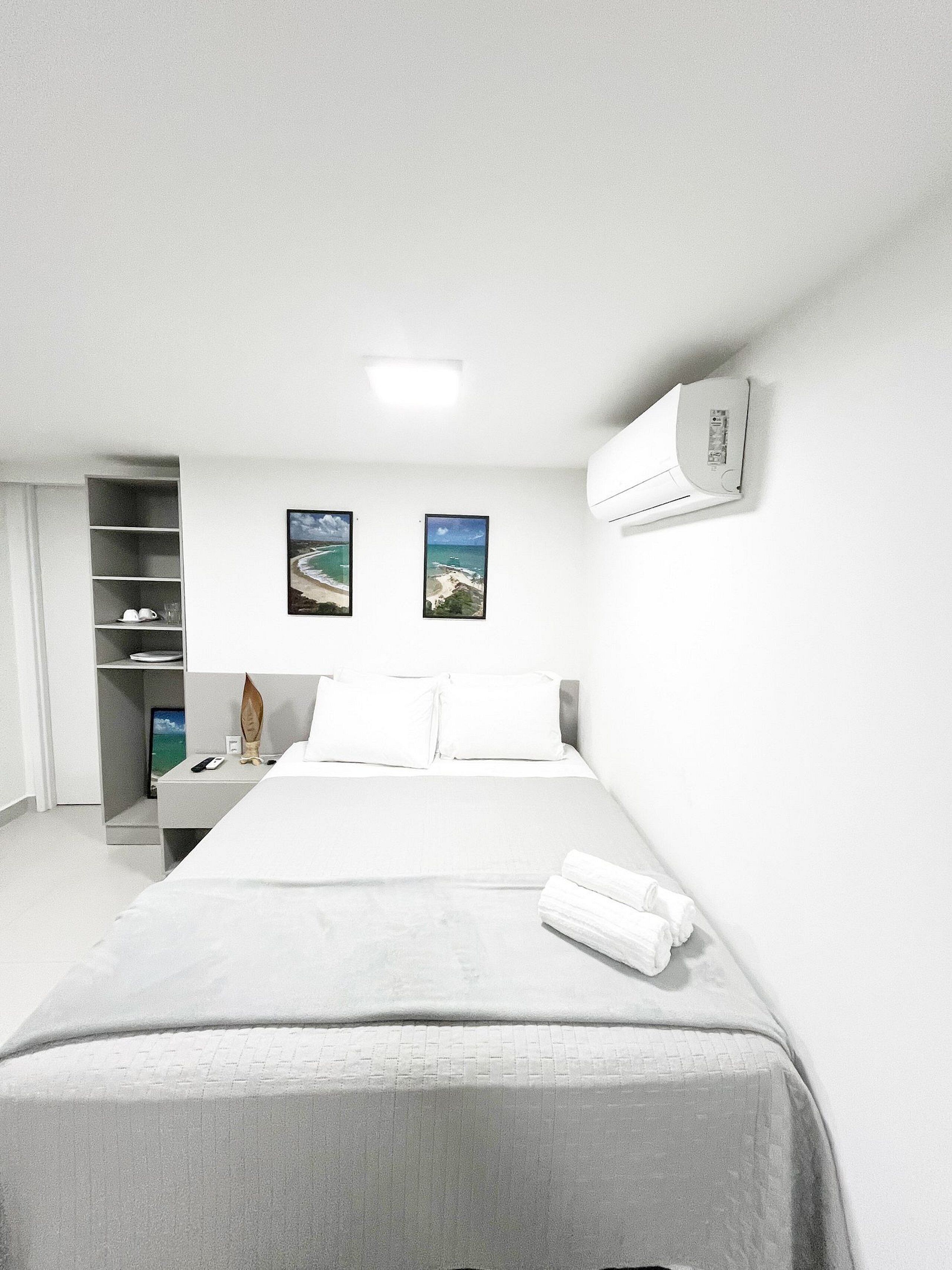 A hotel room with a bed, a flat-screen TV, two framed pictures, a white air conditioner, and a white wardrobe.