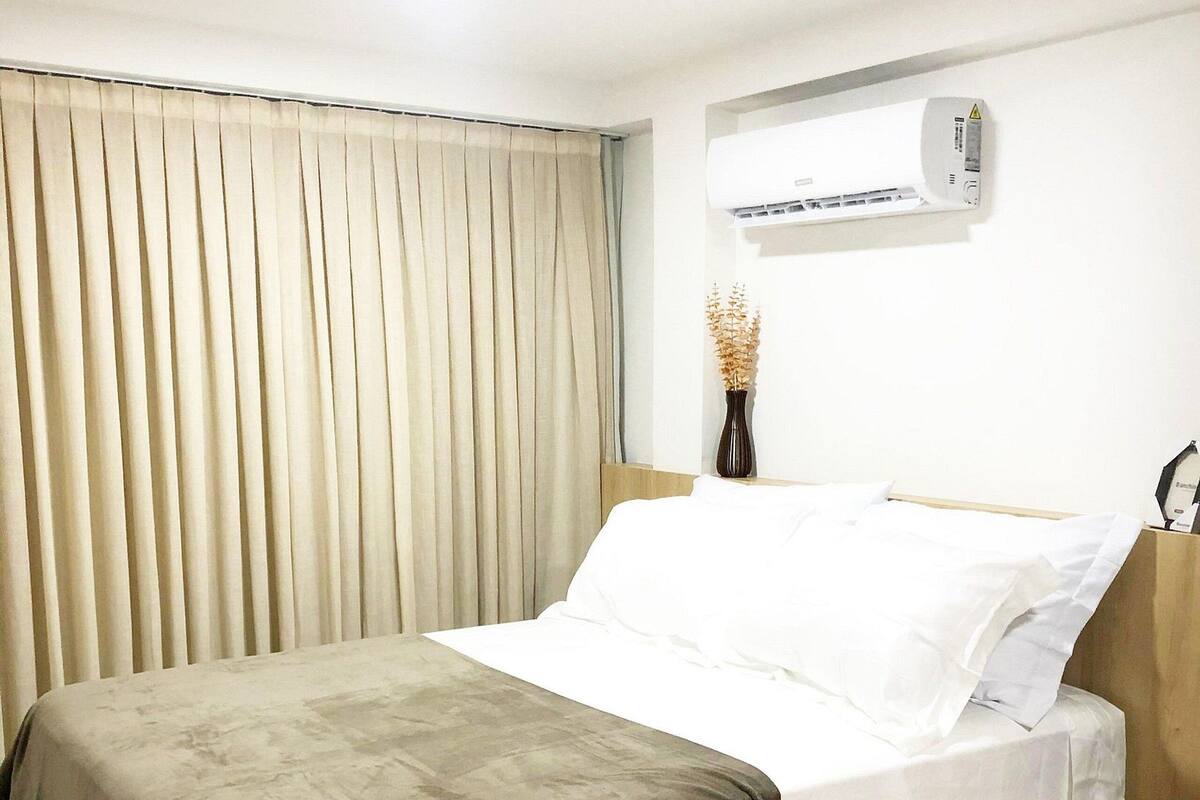 Una cama bien hecha con una manta marrón, sábanas blancas y un jarrón con flores secas en la mesita de noche.