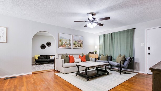 A living room with hardwood flooring, a ceiling fan, a white sofa with colorful cushions, a coffee table, and a green curtain. - River Oaks Retreat- Walkable to Goodfellow AFB (San Angelo)
