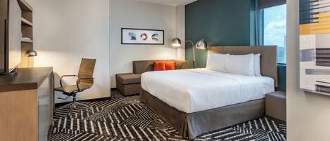 A modern hotel room with a large bed, a desk, a chair, and a desk lamp.