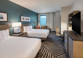 Habitación de hotel con dos camas, un escritorio, un televisor de pantalla plana, un sofá y un amplio ventanal con vistas al exterior.