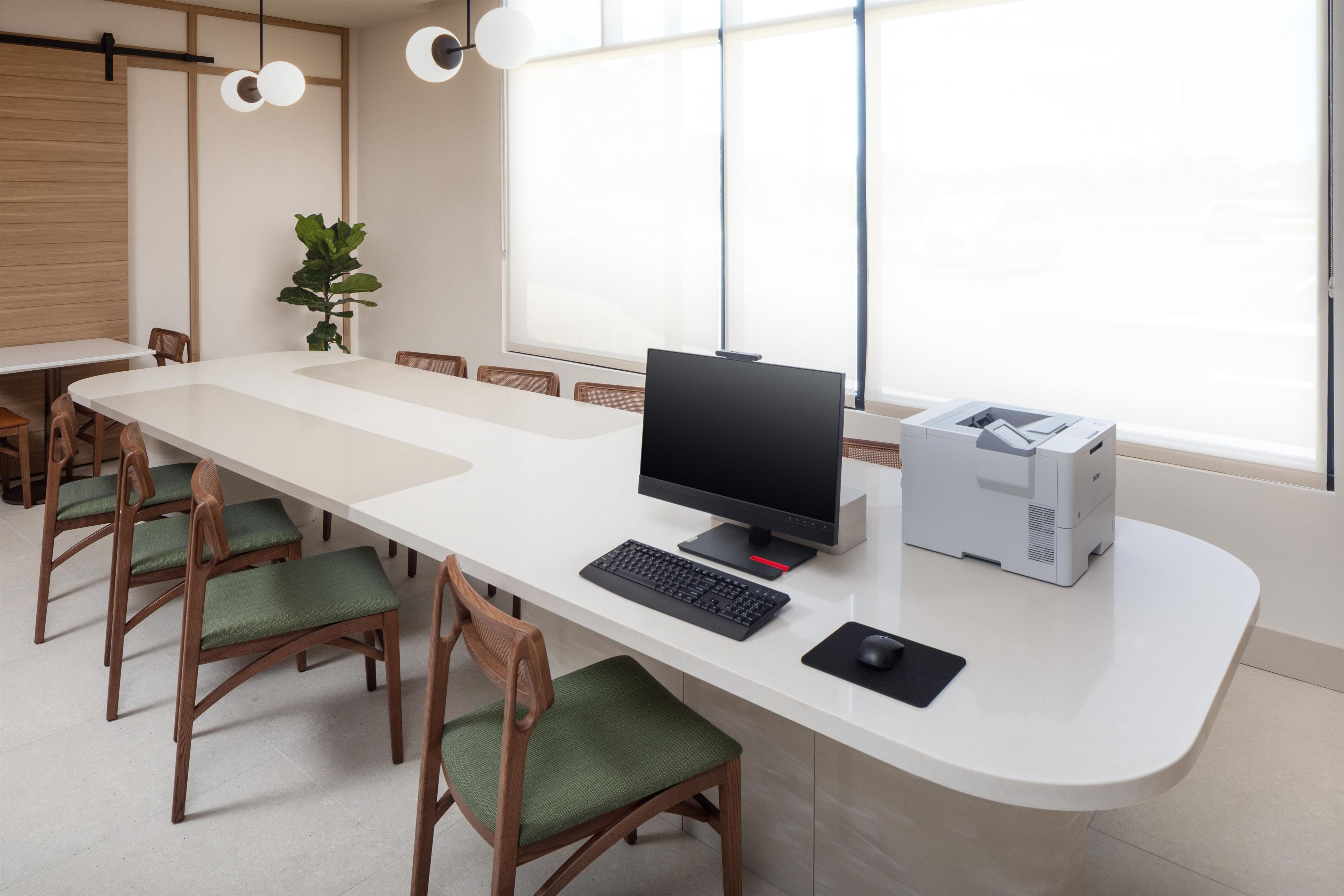 Un espace de bureau moderne avec une longue table blanche, plusieurs chaises en bois dotées de coussins verts, un ordinateur de bureau, une imprimante et une grande fenêtre laissant entrer la lumière naturelle.