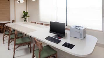 Un espace de bureau moderne avec une longue table blanche, plusieurs chaises en bois dotées de coussins verts, un ordinateur de bureau, une imprimante et une grande fenêtre laissant entrer la lumière naturelle.