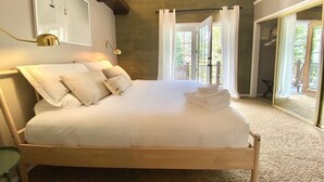 Ein Schlafzimmer mit einem großen Bett, einem Holzkopfende, einem Nachttisch, einem Spiegel und Blick ins Freie durch eine Glastür.