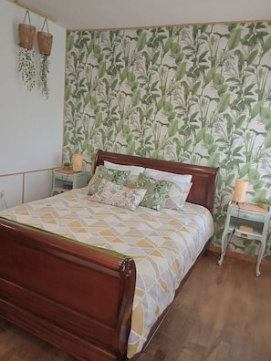 A bedroom with a wooden bed, patterned bedding, a nightstand with a lamp, and a wall with a botanical wallpaper. - Gîte 35kms from Mont Saint Michel (Le Mesnillard)