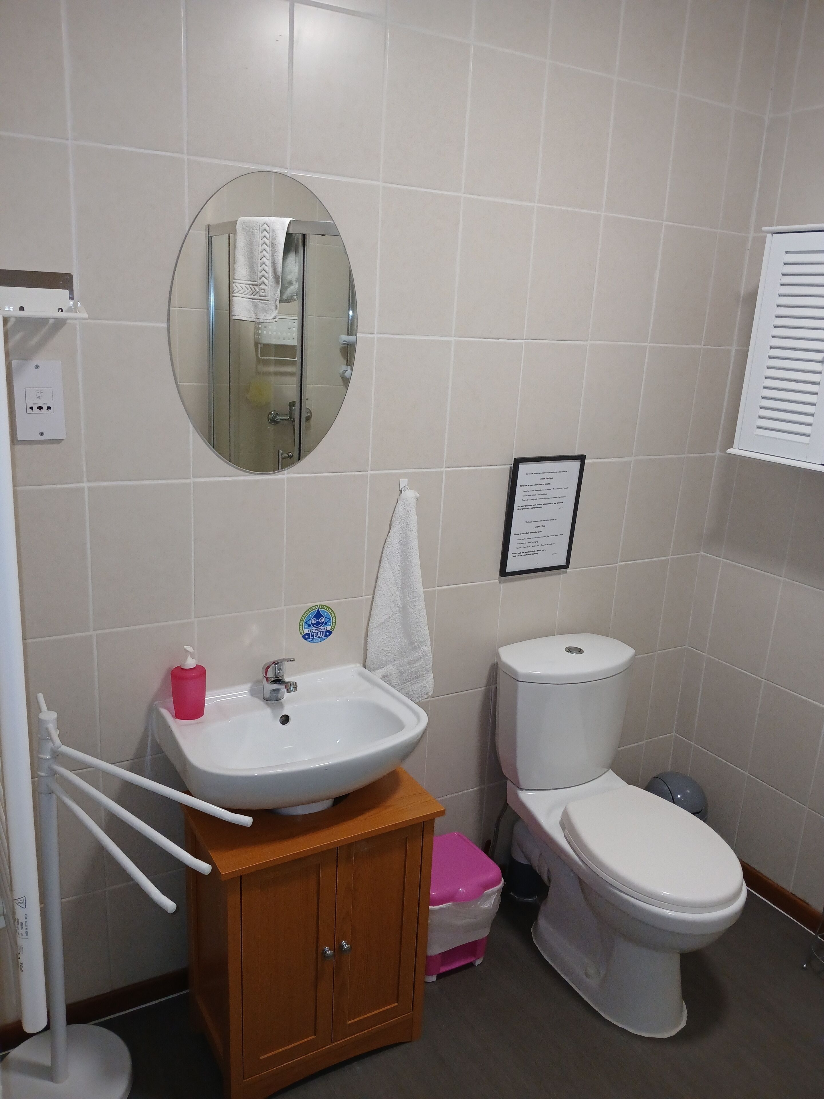 A bathroom with a white sink, mirror, and toilet. There is a wooden cabinet below the sink, a pink trash bin, and a white towel hanging on the wall.
