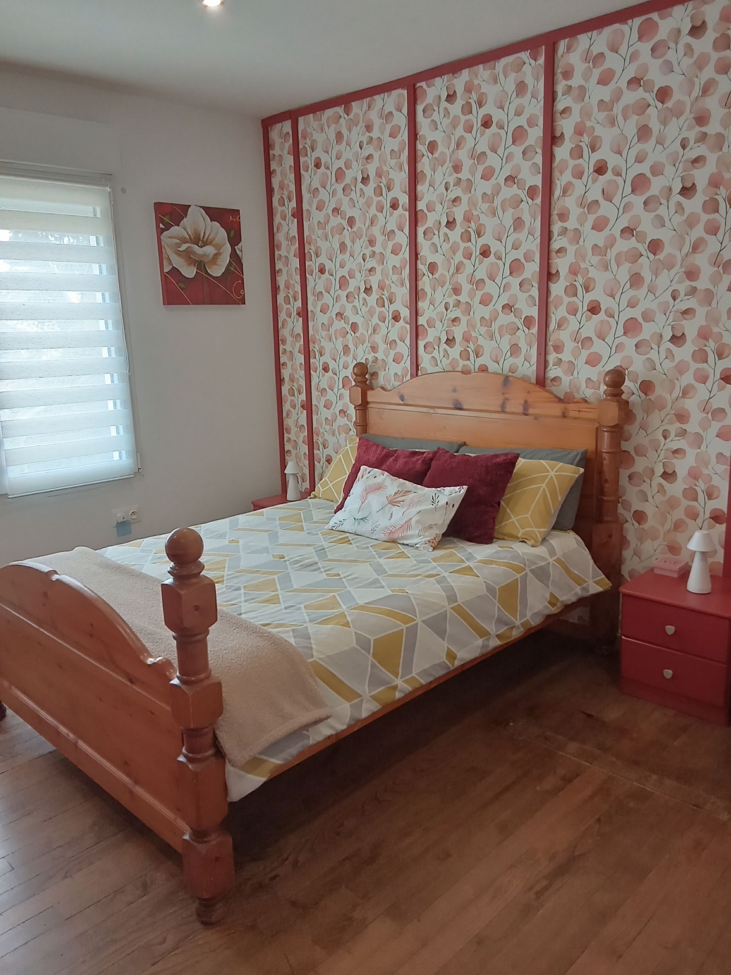 A wooden bed with a geometric patterned bedspread, a red bedside table, and a wall with a floral wallpaper pattern.