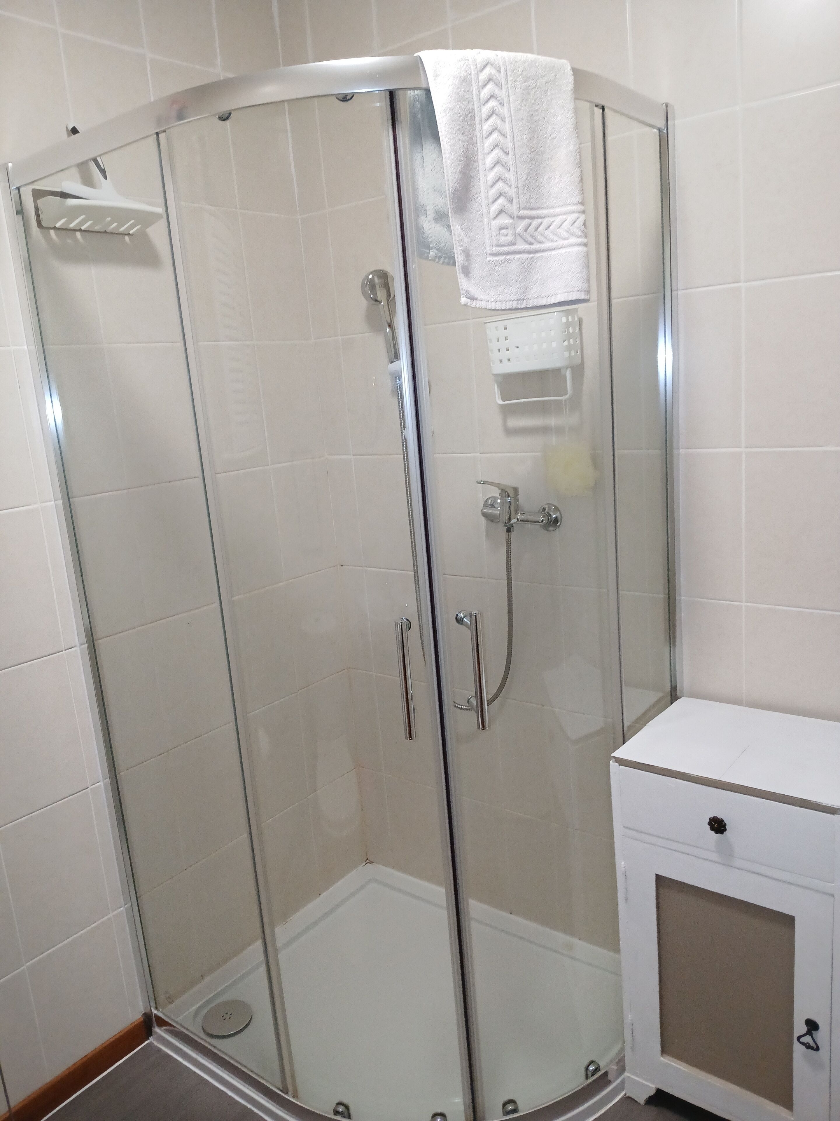 A glass-enclosed shower with a white towel hanging on the door, a white cabinet with a black knob, and a tiled wall.