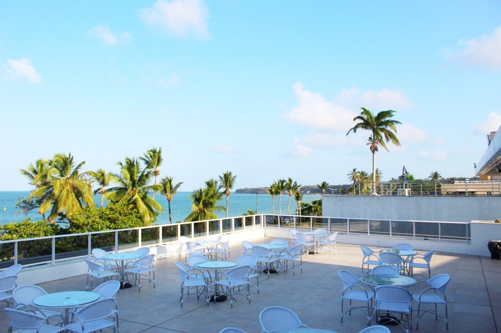 Terraço no telhado com cadeiras e mesas de metal branco, com vista para a costa, palmeiras e o mar.