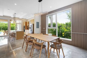A modern kitchen with stainless steel appliances, a dining area with a wooden table and chairs, and large windows offering a view of greenery. - BRAND NEW Villa Between 2 Blues, Beachwalk Villa 5 (Providenciales)