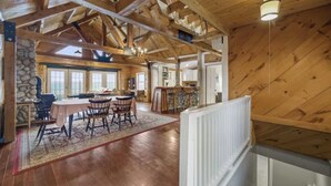 Une salle à manger spacieuse avec un plafond haut et des poutres apparentes, une cheminée en pierre et une vue sur la cuisine en arrière-plan.
