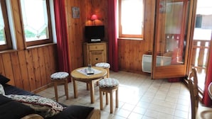 Ein gemütliches Zimmer mit Holzwänden, Fliesenboden, einer Couch, einem kleinen Tisch mit Hocker, einem Fernseher und einer Glastür zum Außenbereich.