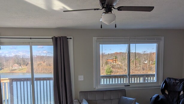 A spacious living room with a gray sofa, a massage chair, a ceiling fan, and a large window offering a view of a lake and a house. - Lakefront Haven w/ 50-ft Dock 4BR Near Punderson Park & Amish Country EV Charger (Newbury)