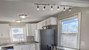 A modern kitchen with white cabinetry, a stainless steel refrigerator, a built-in island with bar stools, and a large window providing natural light. - Lakefront Haven w/ 50-ft Dock 4BR Near Punderson Park & Amish Country EV Charger (Newbury)