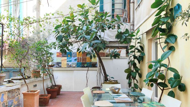 Una mesa con cubierta de vidrio y sillas, rodeada de plantas en macetas y una pared de azulejos.