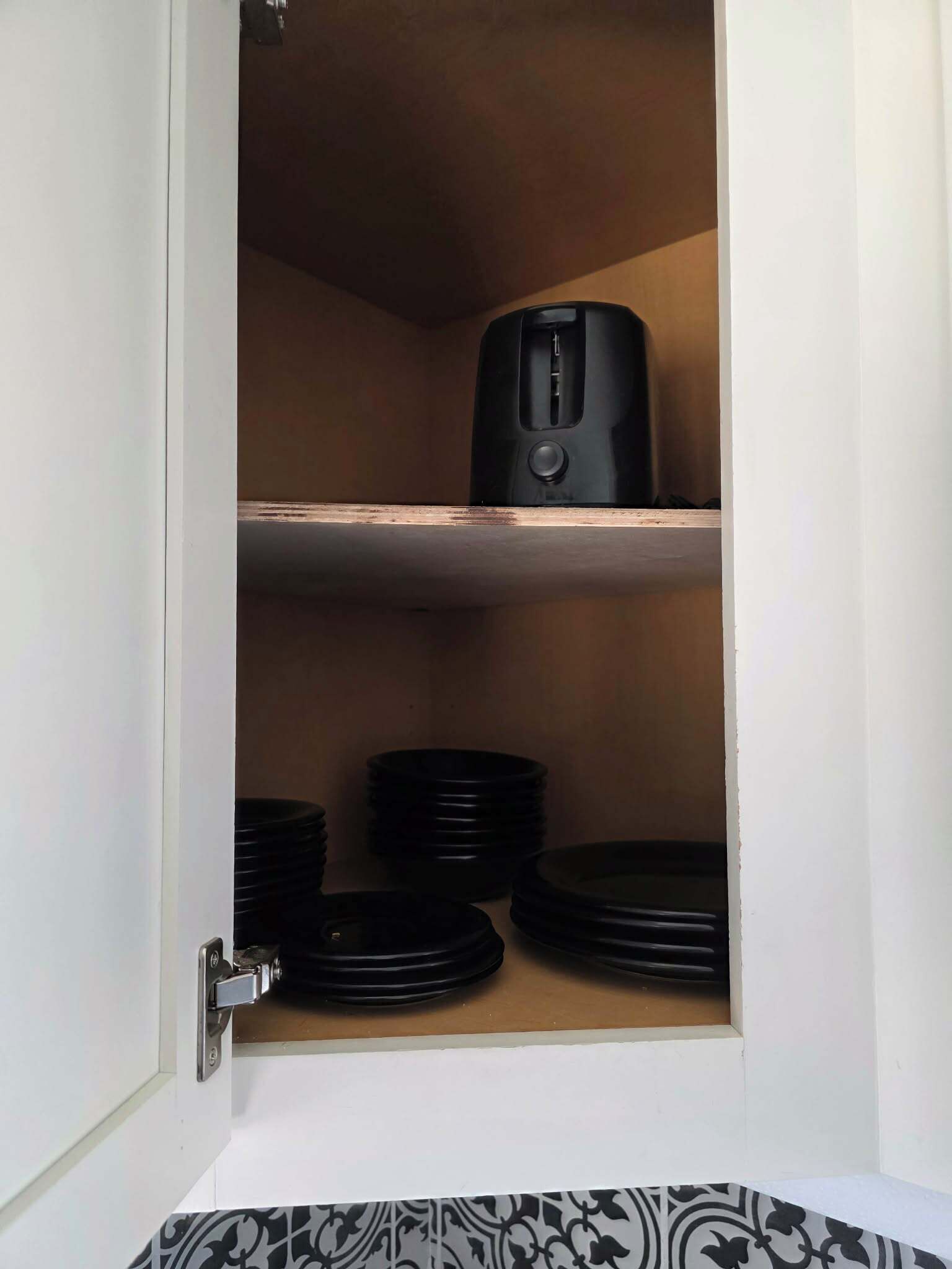 A cabinet with a toaster on the top shelf and black plates stacked on the bottom shelf.