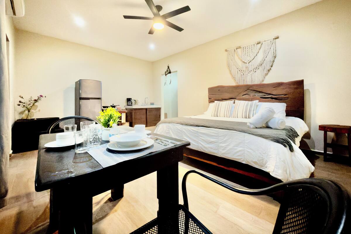 Una habitación de hotel con cama, una mesa de comedor con sillas, kitchenette y ventilador de techo.