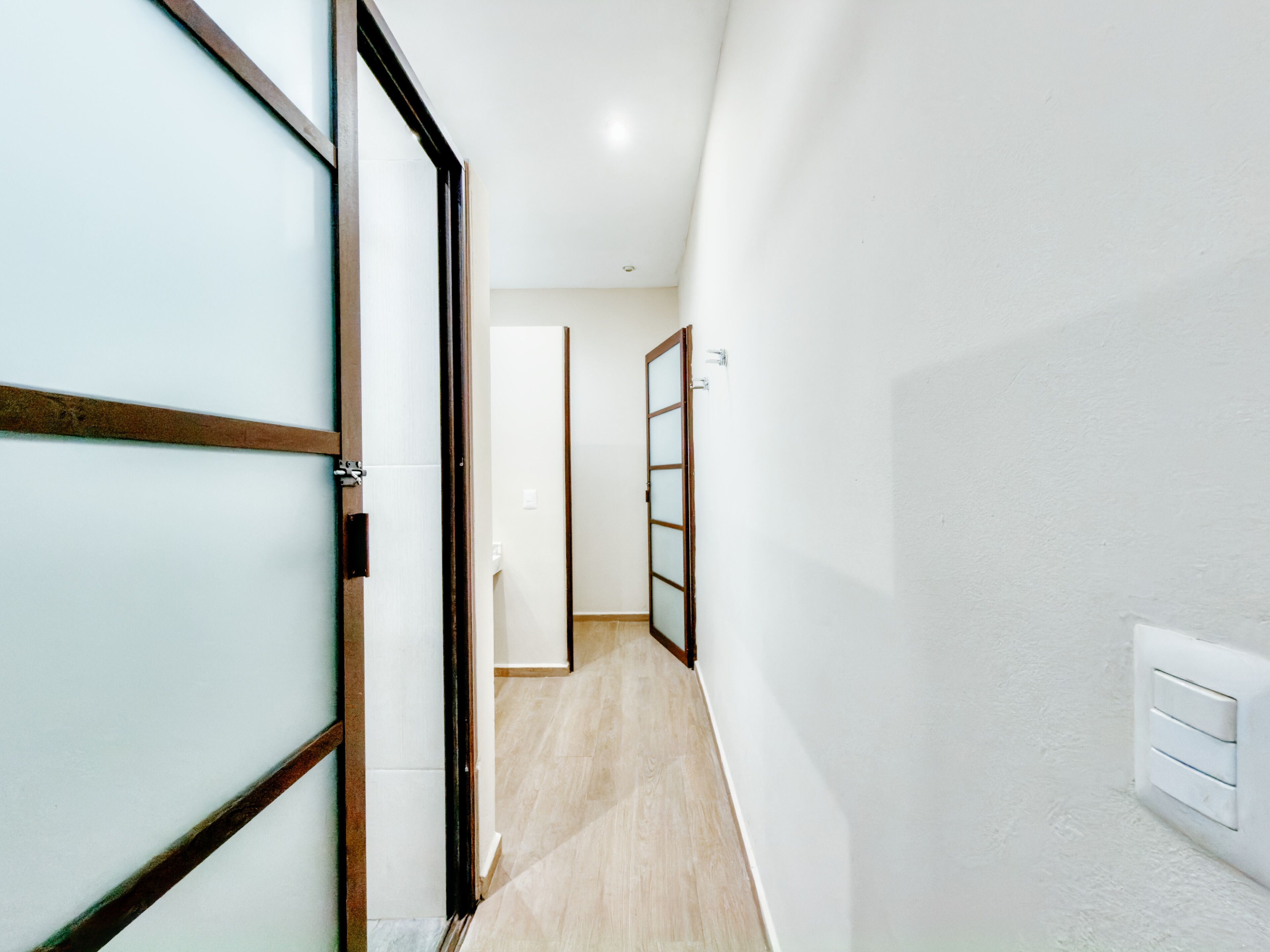Un pasillo con piso de madera, paredes blancas y una puerta corredera de vidrio a la izquierda.