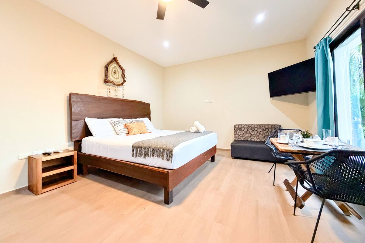 Habitación de hotel con cama de madera, una mesita de noche pequeña, un sofá, una mesa de comedor con sillas y un ventilador de techo.