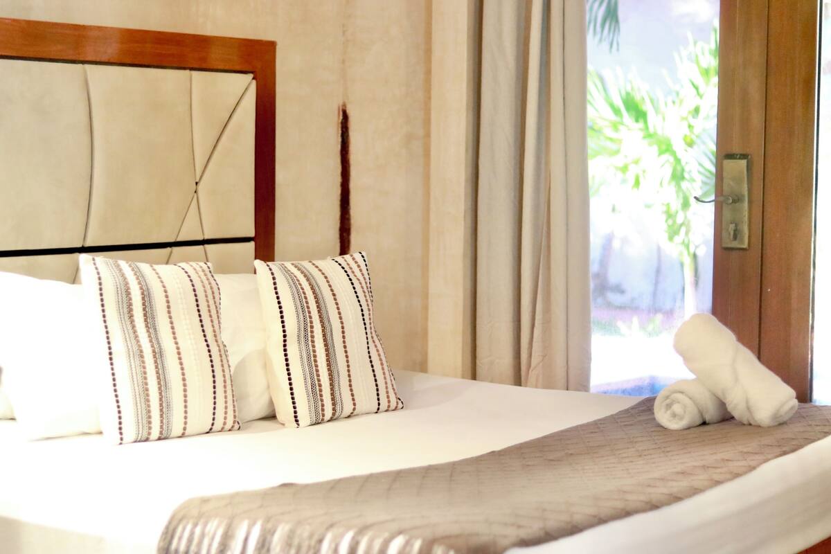Habitación de hotel con cama, almohadas, una colcha, ventana con cortinas y una toalla sobre la cama.