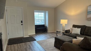 A living room with a dark brown leather couch, a small side table with a lamp, a bench, and a door with a doorknob. - Kozy Keizer Getaway with Fireplace (Keizer)