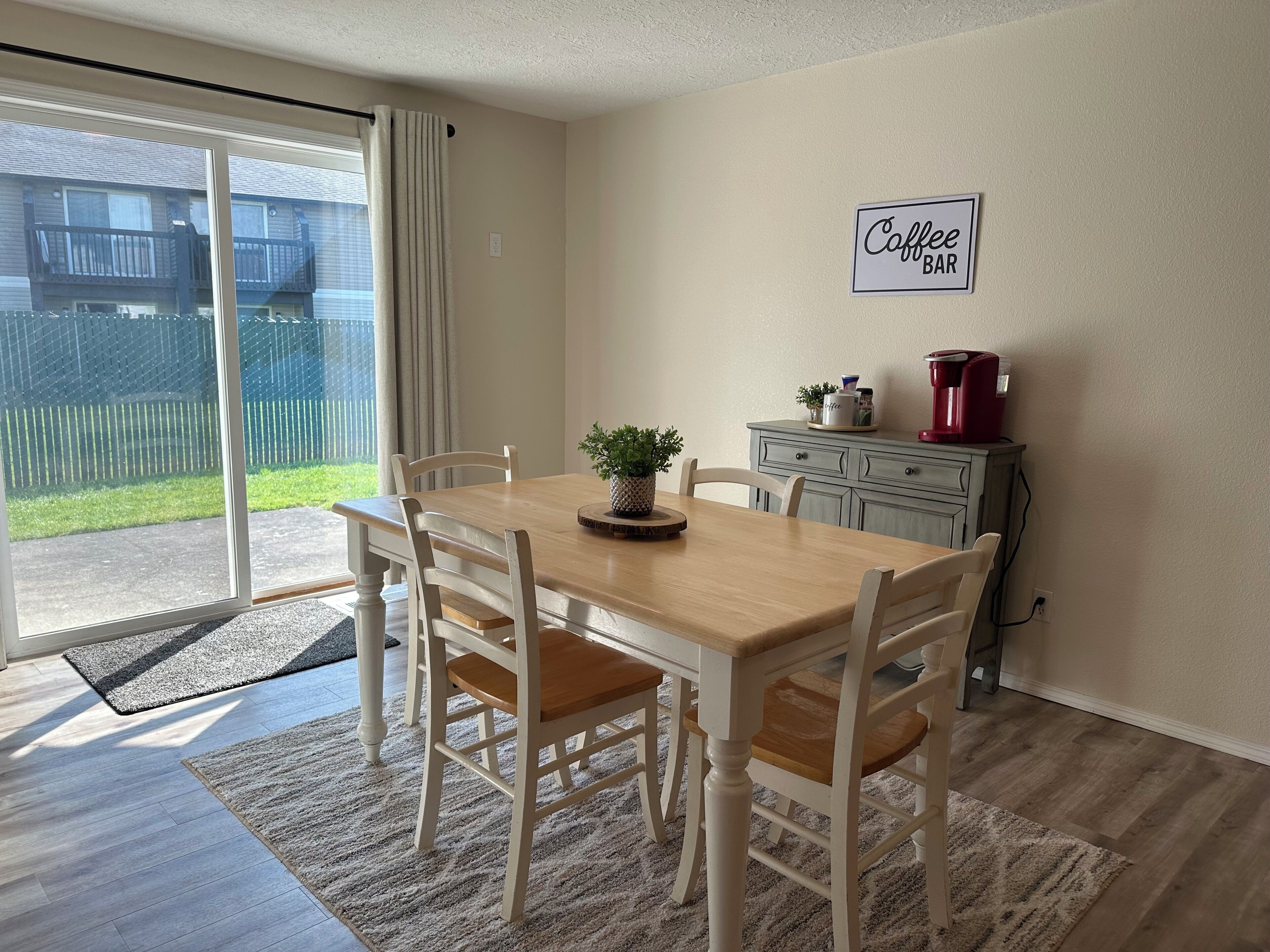 Un comedor con una mesa y sillas de madera, un letrero que indica cafetería y vista a un patio trasero cercado.