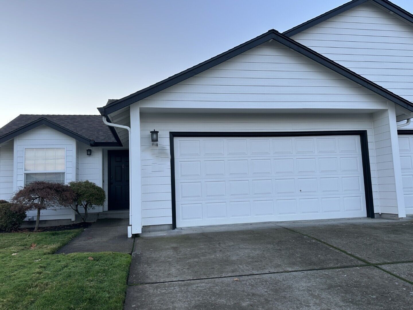 A single-story house with a white exterior, a garage with a black door, and a small front yard with a bush.