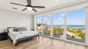 Ein Schlafzimmer mit einem Bett, einem Nachttisch, einem Deckenventilator und großen Fenstern mit Blick auf Strand und Meer.