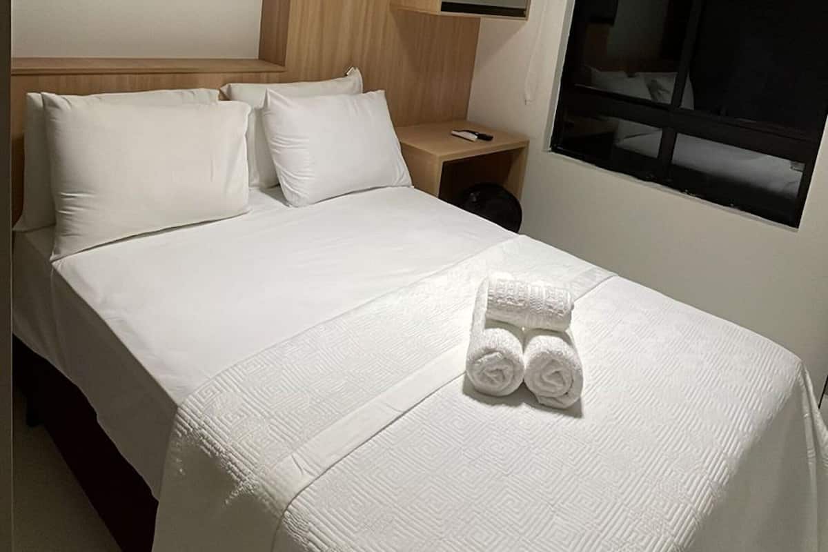 Una cama bien hecha con sábanas blancas y dos toallas enrolladas, una mesita de noche con una lámpara, y una ventana con vistas al exterior.