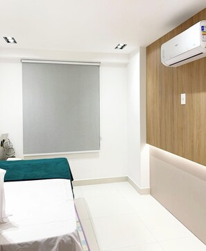 Quarto de hotel com cama, janela com persiana, ar condicionado e parede de destaque em madeira.