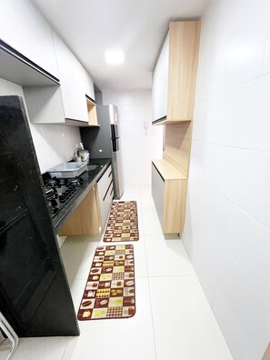 Eine schmale Küche mit weißen Schränken, einer schwarzen Arbeitsplatte und einem Kühlschrank. Auf dem Boden liegen zwei Teppiche mit Muster.