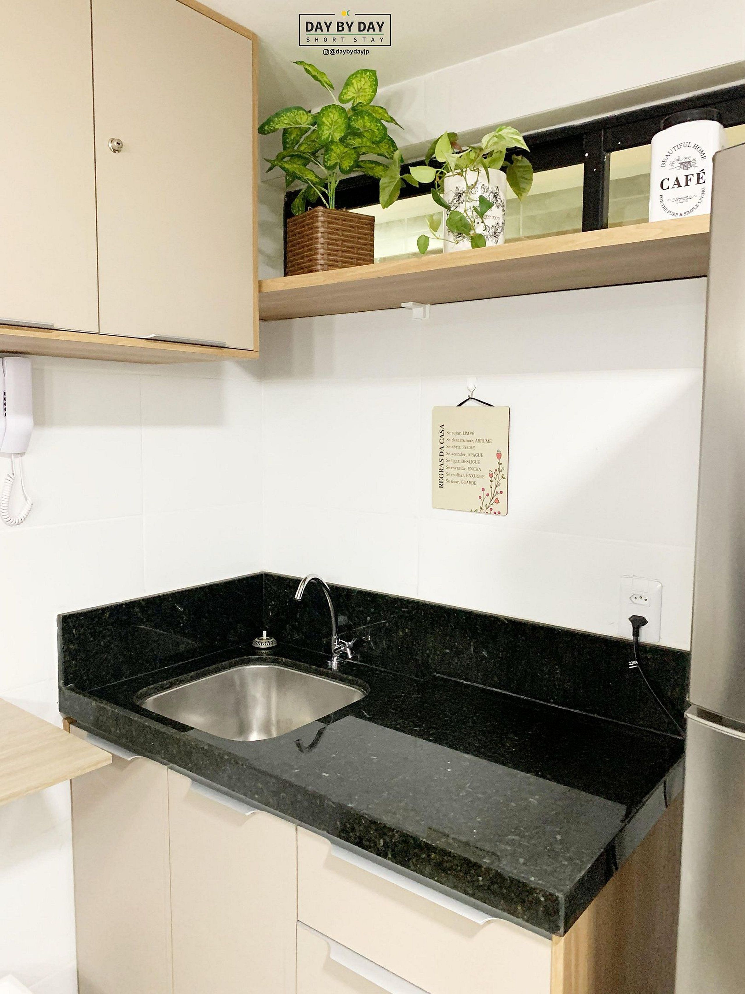 A kitchen with a black granite countertop, a stainless steel sink, and a wooden shelf above holding plants and a sign.