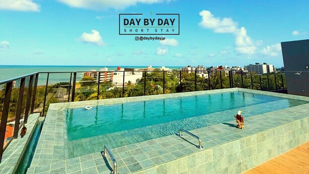 A rooftop infinity pool with clear blue water, surrounded by a wooden deck and glass railings, overlooking a cityscape and the ocean. - Dutra Ocean Flat By DayByDay (Joao Pessoa)