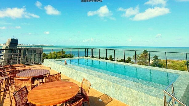 A rooftop pool area with a clear view of the ocean, wooden tables and chairs, and a glass balustrade. - Dutra Ocean Flat By DayByDay (Joao Pessoa)