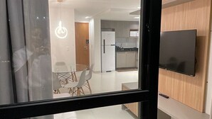 A modern living room with a sofa, a dining area with chairs, a kitchen with a refrigerator, and a TV mounted on the wall. - Dutra Ocean Flat By DayByDay (Joao Pessoa)