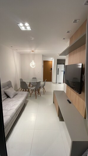 A modern living room with a sofa, a dining table with chairs, a TV mounted on a wooden panel, and recessed ceiling lights.