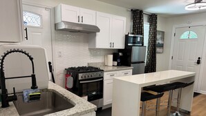 Una cocina moderna con armarios blancos, una campana extractora de acero inoxidable, una estufa a gas, un microondas y un refrigerador. Hay una zona de bar con taburetes.