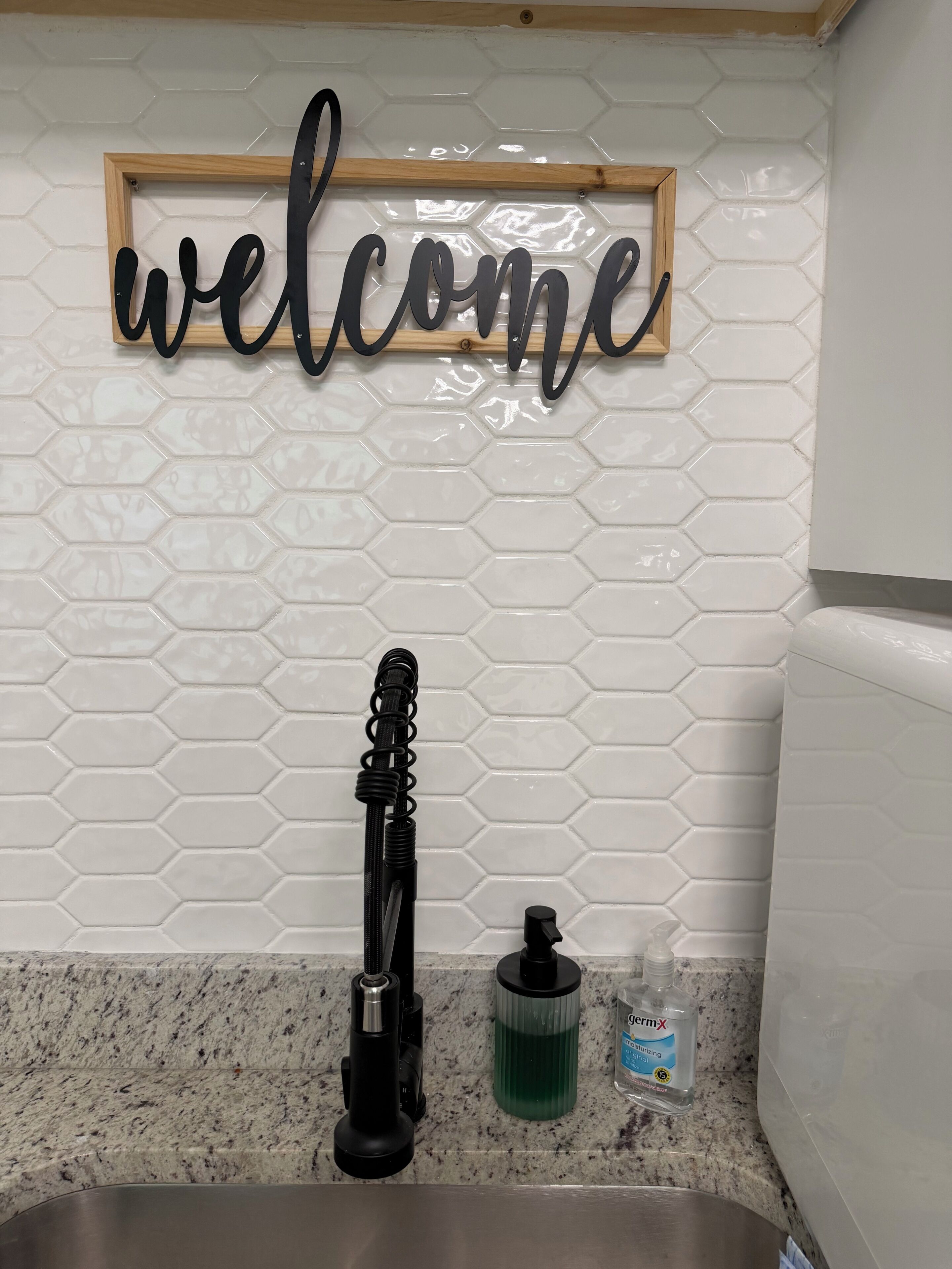 Una cocina con un cartel que dice 'welcome', un lavamanos, un grifo, un dispensador de jabón y una botella de jabón de manos.