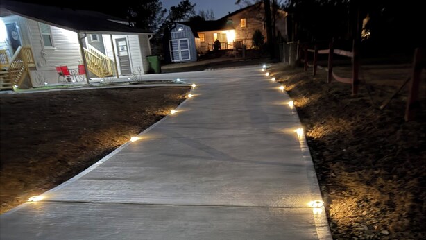 Un camino de concreto con luces regularmente espaciadas, bordeado por una valla de madera y un cobertizo pequeño.