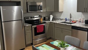 Een moderne keuken met grijze kastjes, roestvrijstalen apparatuur, een houten eettafel met placemats, en een rode handdoek aan de oven.
